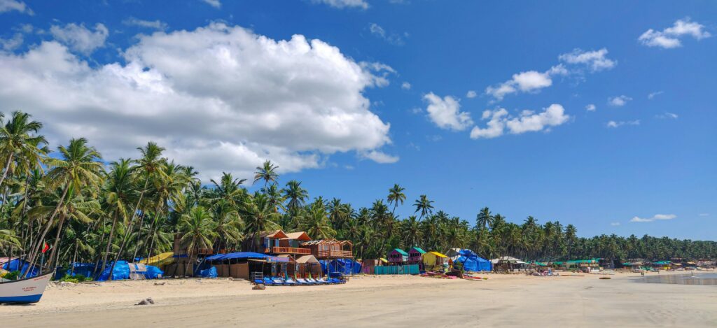 Panoramic view of a tropical beach with palm trees and vibrant beach huts under a clear blue sky.